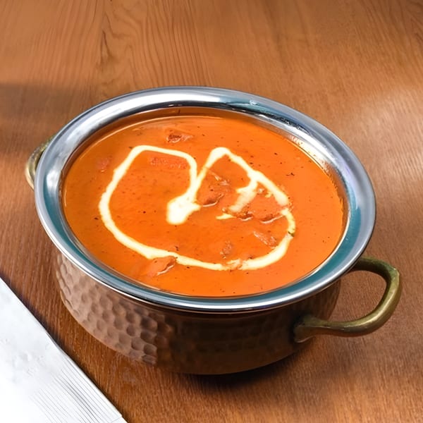A copper karahi bowl filled with orange Indian curry, garnished with cream swirls, on a wooden table.
