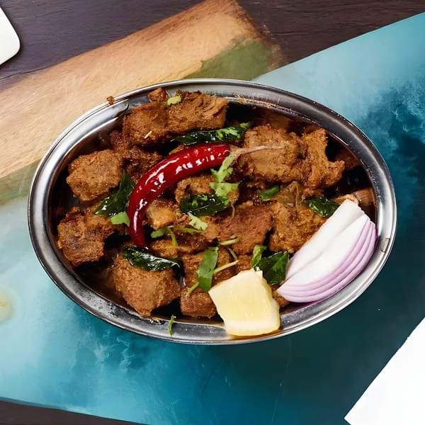 A metal bowl of Indian meat curry, garnished with red chili, cilantro, onion, and lemon.