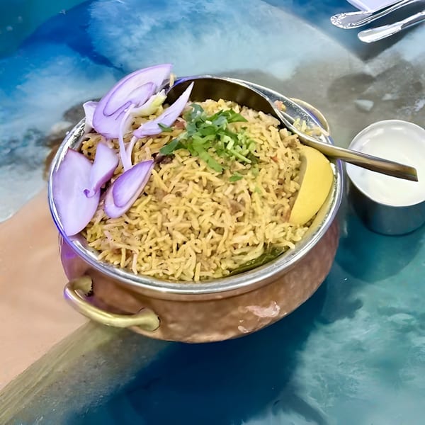 A copper pot of Indian biryani rice with red onions, cilantro, lemon, and a side of raita.